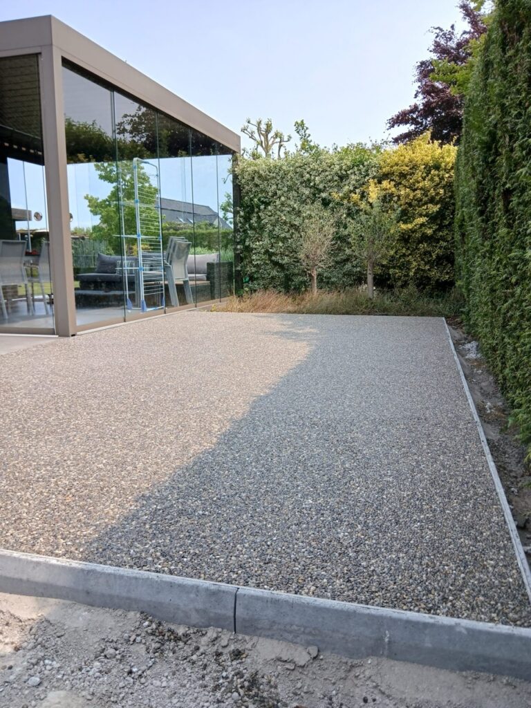 Strak aangelegd Terradec en Vuba steentapijt terras naast glazen tuinkamer in groene tuin door Xtraverde Poperinge